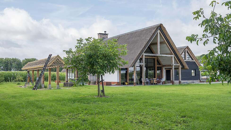 Een modern landelijk huis met rieten dak en zwarte gevelbekleding, omgeven door een groene tuin met notenbomen en een natuurvijver